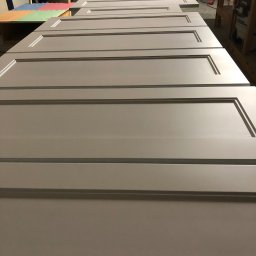 MIĘDZY SĘKAMI JAKUB WIERNICKI-BALCER - Szare, gotowe fronty meblowe z frezowanymi ramkami leżące obok siebie w warsztacie stolarskim, przygotowane do montażu. Widoczne tło warsztatowe.