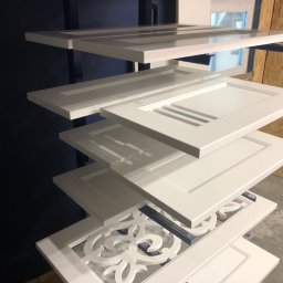 MIĘDZY SĘKAMI JAKUB WIERNICKI-BALCER - Białe, lakierowane fronty meblowe z różnymi wzorami, ułożone warstwowo na metalowym stojaku w lakierni. Widoczne detale wykończenia i struktura drewna.