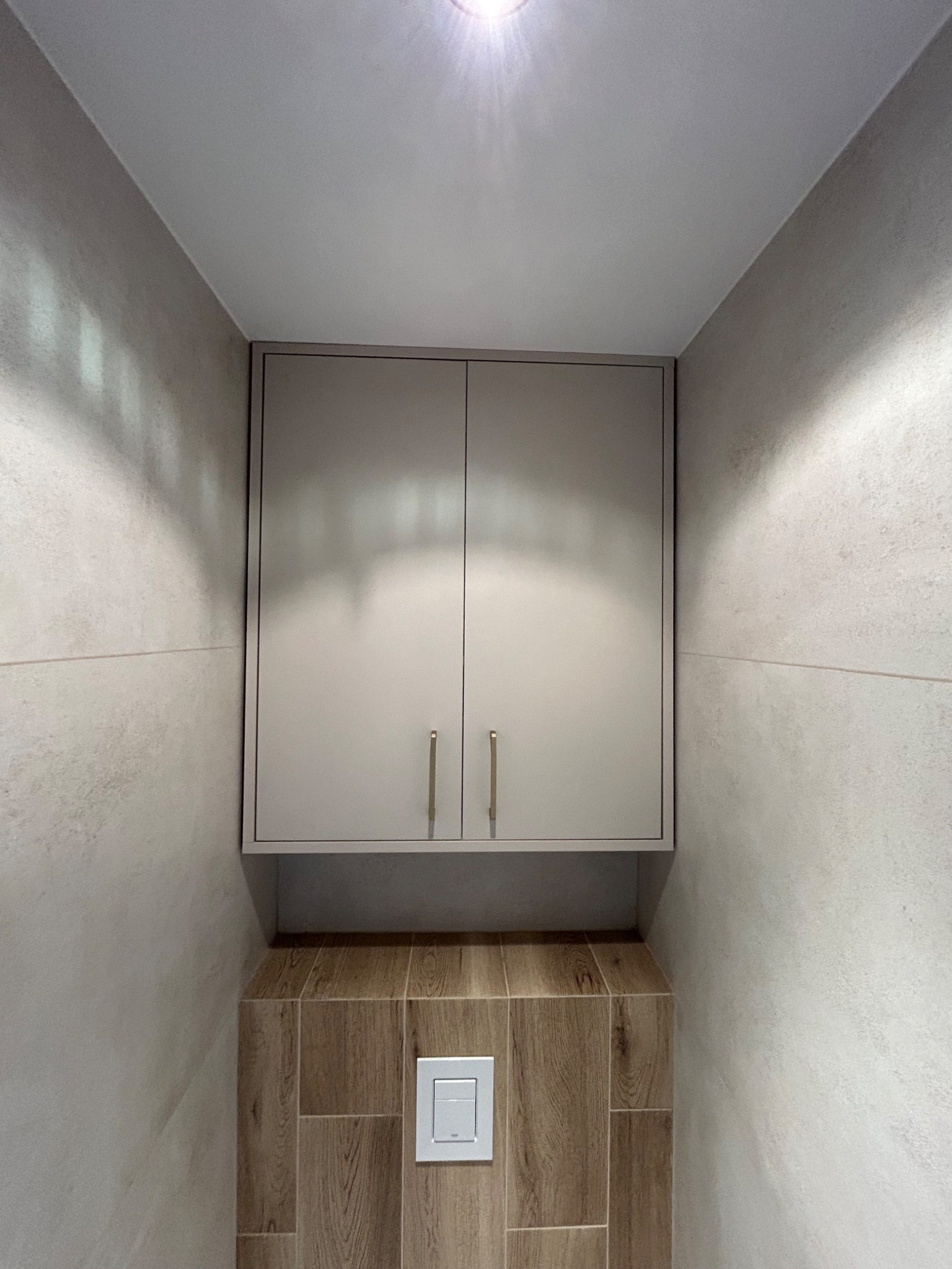 Zamknięta szafka wisząca w łazience, z dwoma drzwiczkami i długimi uchwytami, nad zabudową toalety z brązowymi płytkami imitującymi drewno.