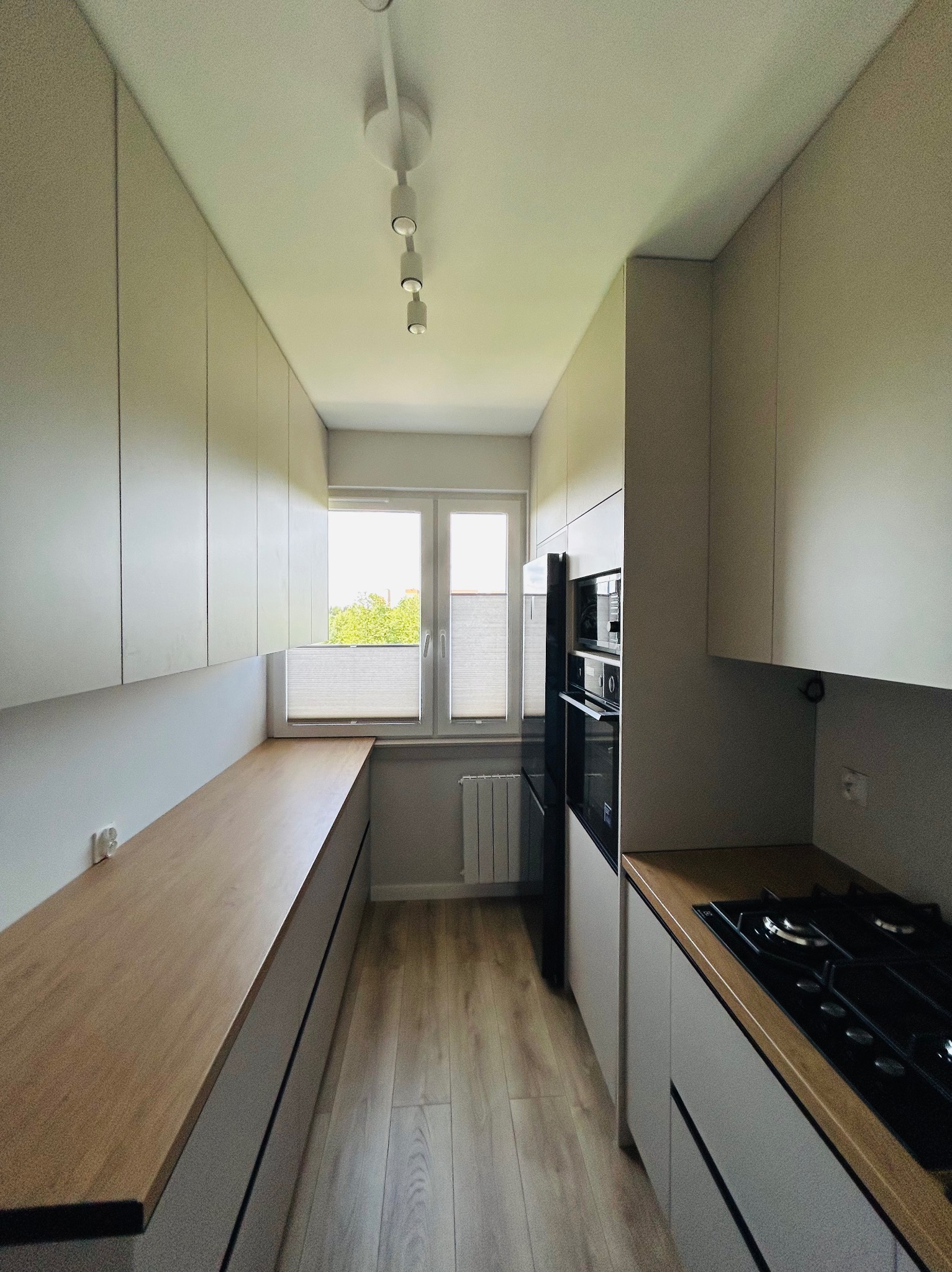 Wąska kuchnia z zabudową po obu stronach, jasne fronty szafek i drewniany blat roboczy. Widoczne okno z roletami plisowanymi i sprzęt AGD w zabudowie.