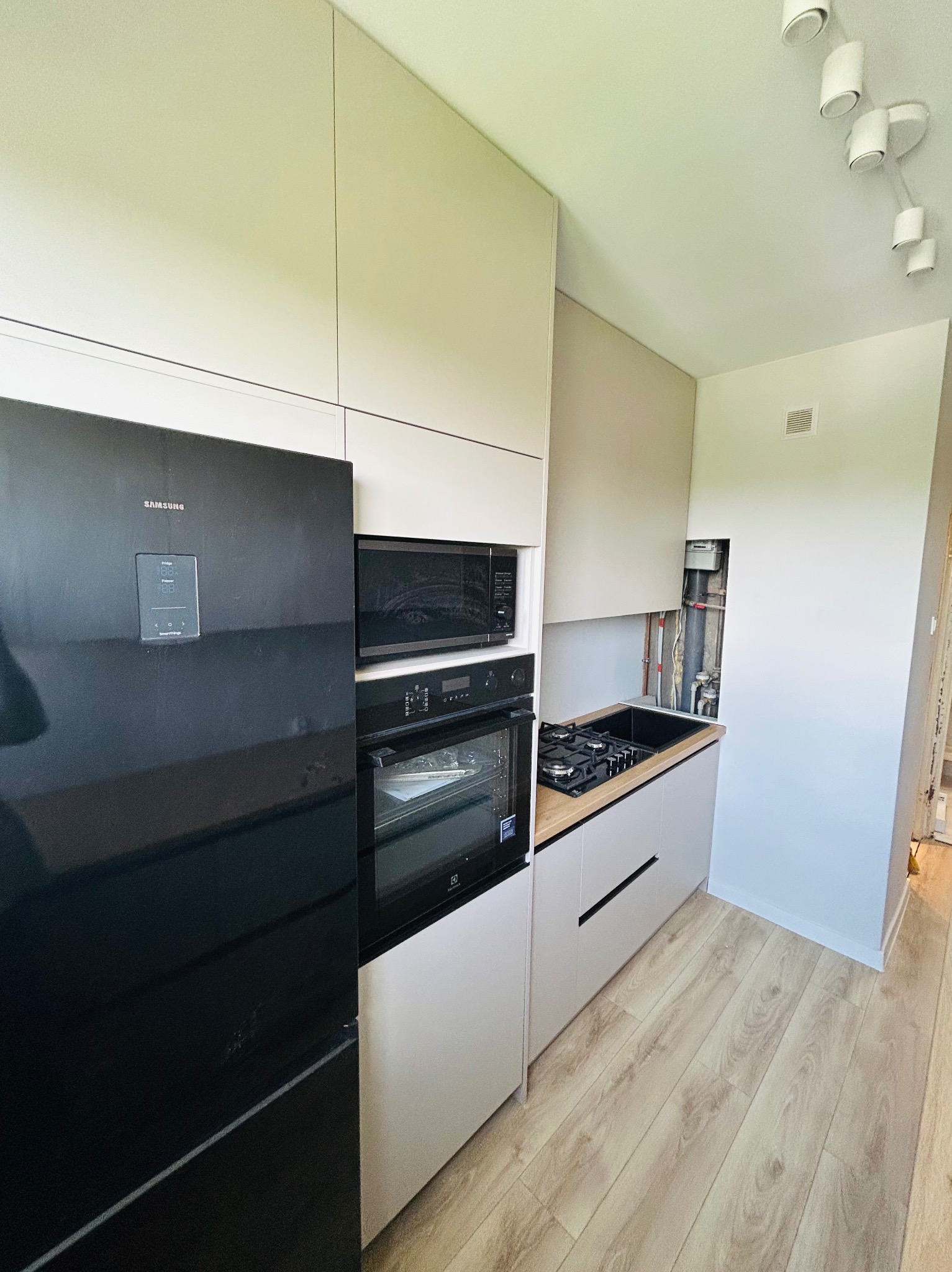 Nowoczesna kuchnia w odcieniach beżu i bieli w Wygodzie. Zabudowa kuchenna z czarnym sprzętem AGD i drewnianym blatem. Widoczne piony instalacyjne.