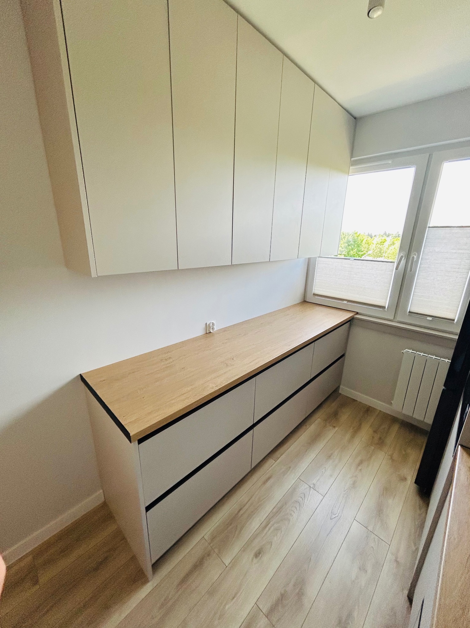 Jasna zabudowa meblowa z drewnianym blatem przy oknie, z górnymi szafkami i dolnymi szufladami z czarnymi akcentami, na podłodze panele imitujące drewno.