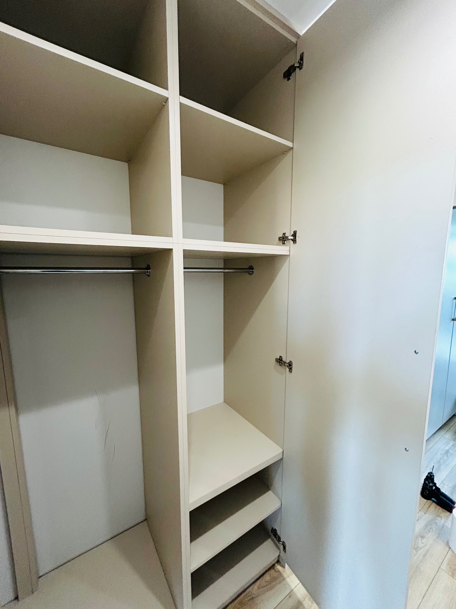 Jasna szafa na wymiar z otwartymi drzwiami, widoczne półki i drążki. Dolne półki na buty. Na podłodze leży wiertarka. Minimalistyczny design.