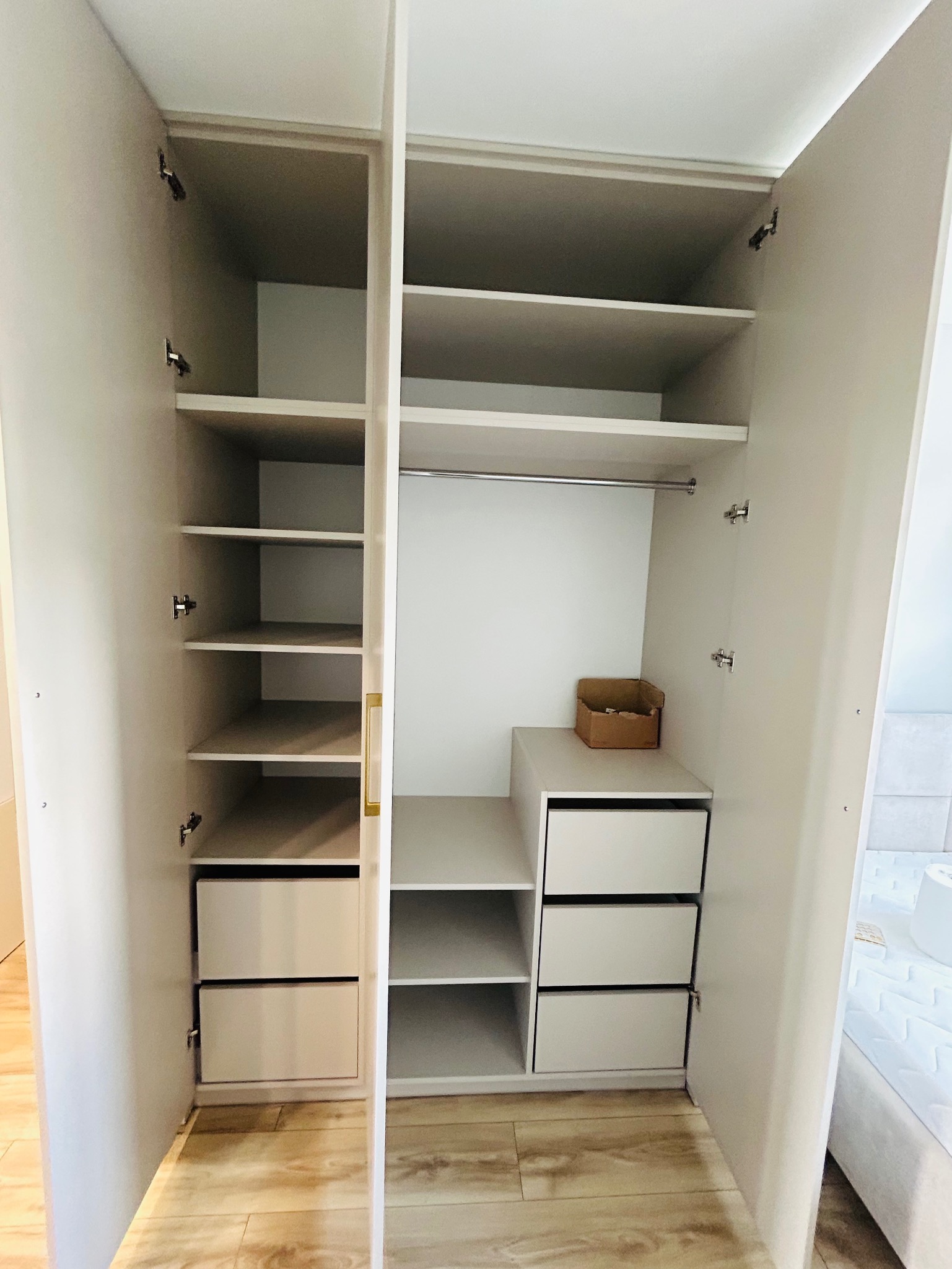 Jasna szafa na wymiar z otwartymi drzwiami, ukazująca półki i szuflady. Widoczna kartonowa skrzynka na jednej z półek. Ujęcie z perspektywy wnętrza pomieszczenia.