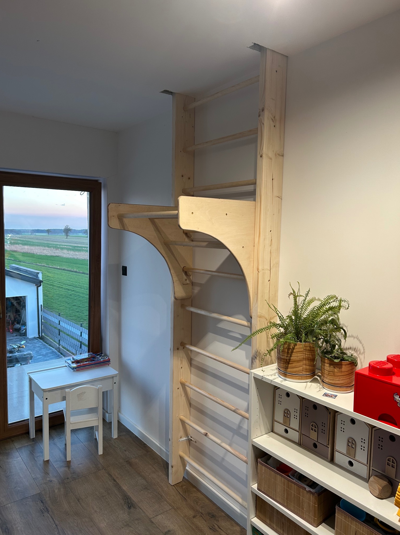 Drewniana drabinka gimnastyczna w pokoju dziecięcym, zamontowana przy ścianie, obok okna z widokiem na zewnątrz i biurka z krzesełkiem. Obok regał z dekoracjami.