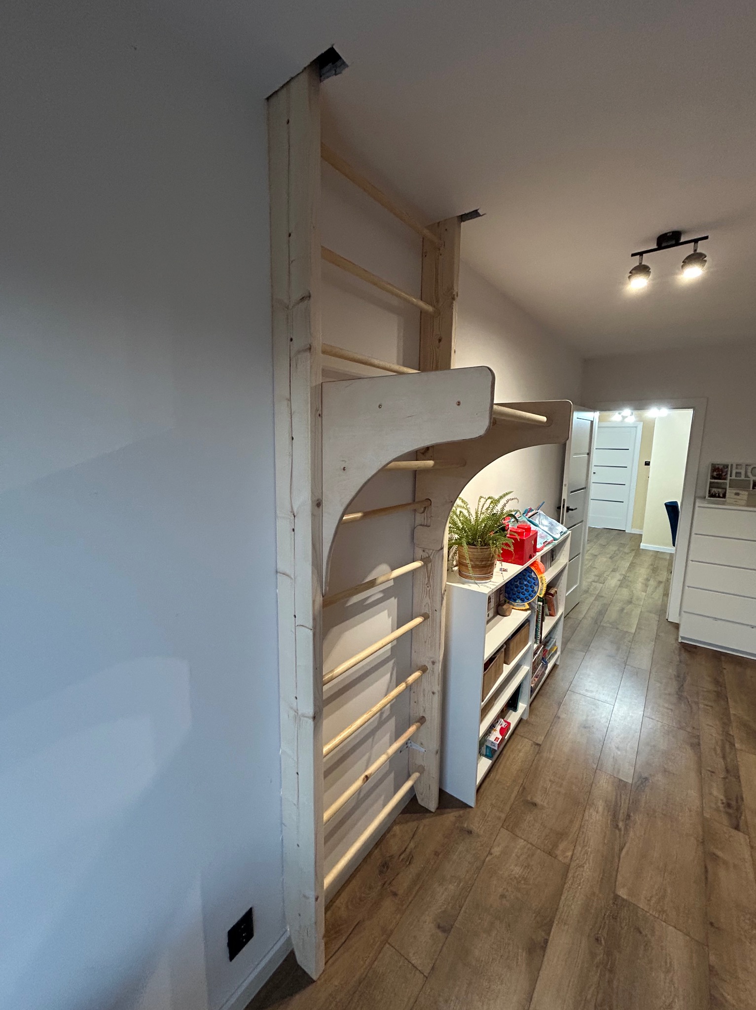 Drewniana drabinka gimnastyczna, mocowana do ściany i sufitu, z dodatkowymi elementami do ćwiczeń, ustawiona w pokoju dziecięcym. Widoczne półki z zabawkami.