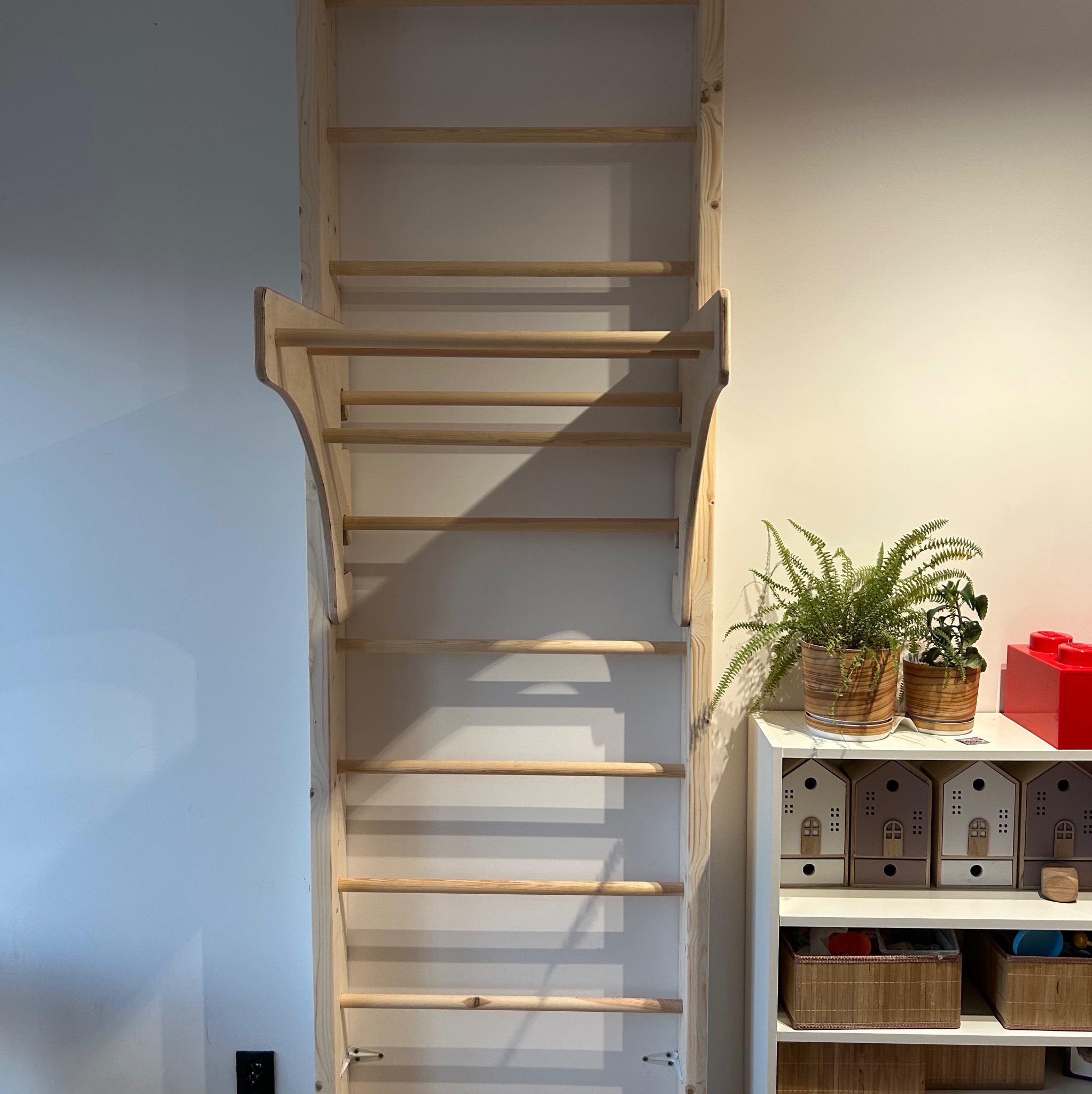 Drewniana drabinka gimnastyczna dla dzieci, zamontowana na białej ścianie. Obok biały regał z dekoracjami i roślinami. Jasne drewno, minimalistyczny design.