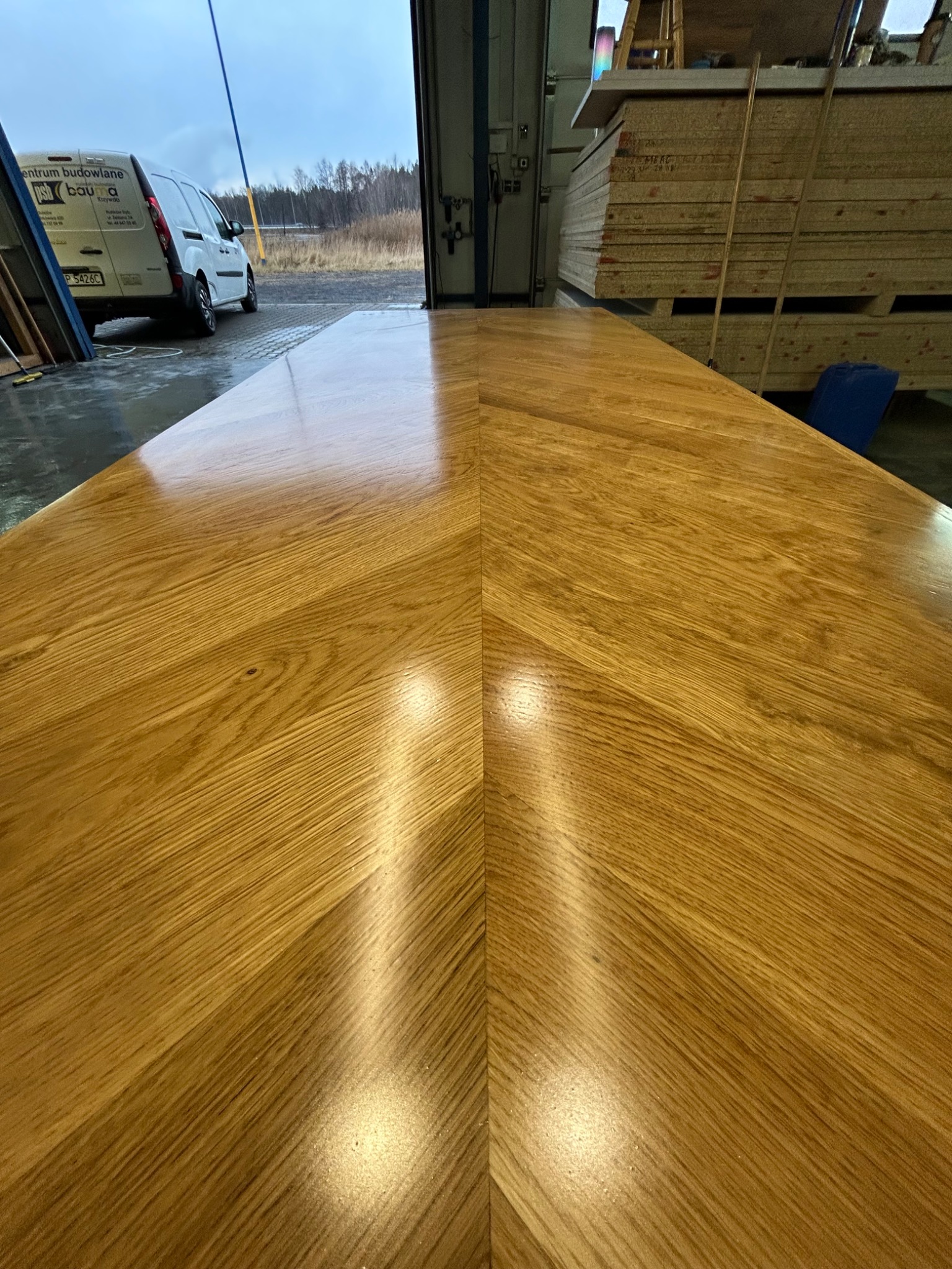 Polerowany blat z drewna o unikalnym wzorze słojów, z odbiciami światła, na tle hali produkcyjnej z zapasem materiałów i samochodem dostawczym.