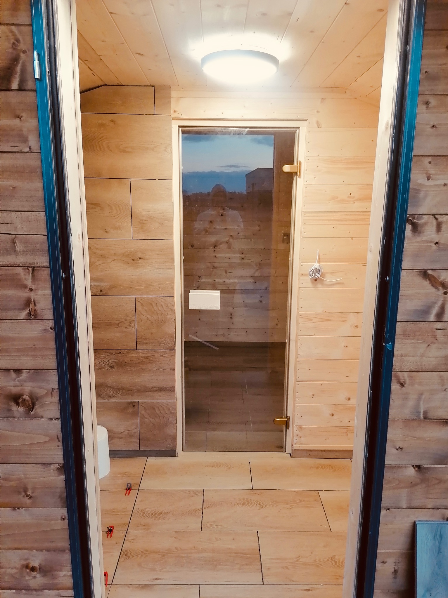 Wnętrze sauny z drewnianym wykończeniem ścian i podłogi z płytek imitujących drewno. Widoczna szklana tafla drzwi. Ujęcie z perspektywy wejścia.