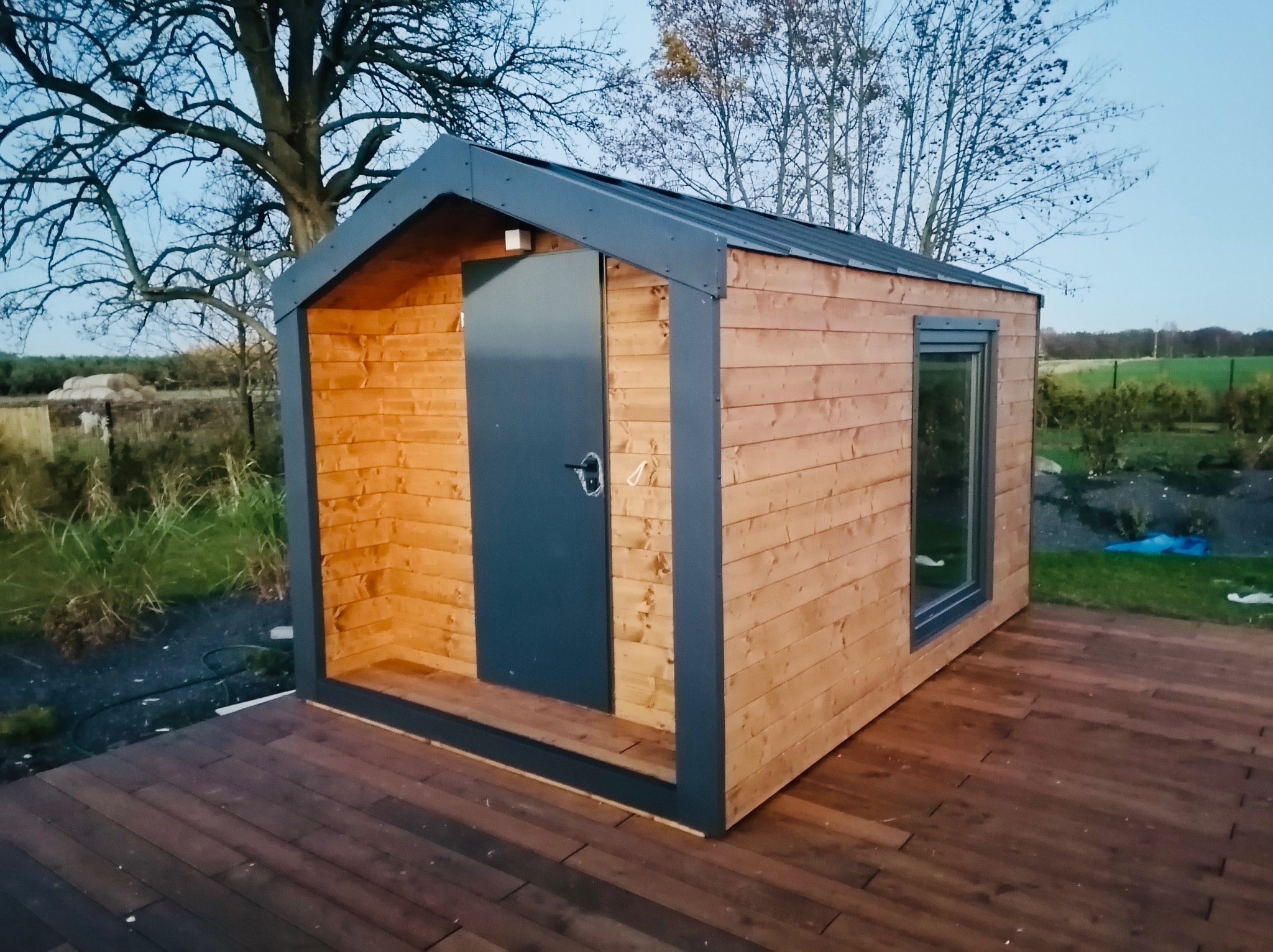 Nowoczesna, drewniana altana z szarymi elementami wykończeniowymi, umieszczona na drewnianym tarasie. W tle widoczne drzewa i pole.