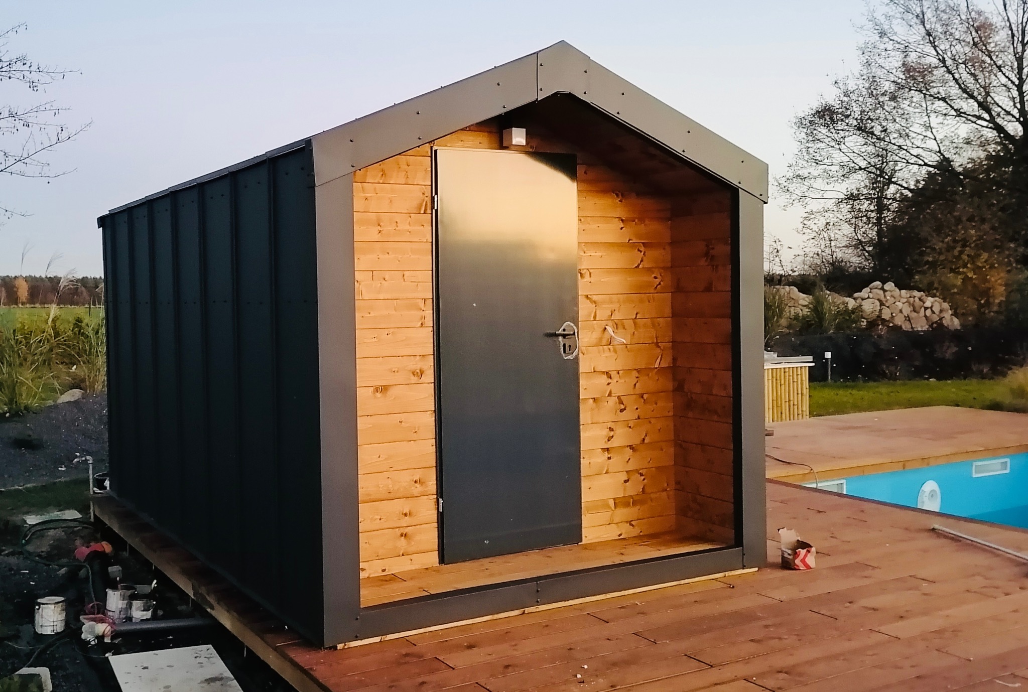 Nowoczesna, drewniana altana z metalowym dachem i drzwiami, umieszczona na tarasie z widokiem na basen i zieleń. Widoczne narzędzia i materiały budowlane.