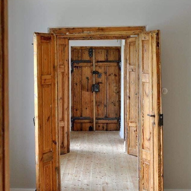 Otwarta, drewniana rama drzwiowa z widokiem na zamknięte, masywne drzwi z metalowymi okuciami. Widoczna podłoga z jasnego drewna i szara ściana.