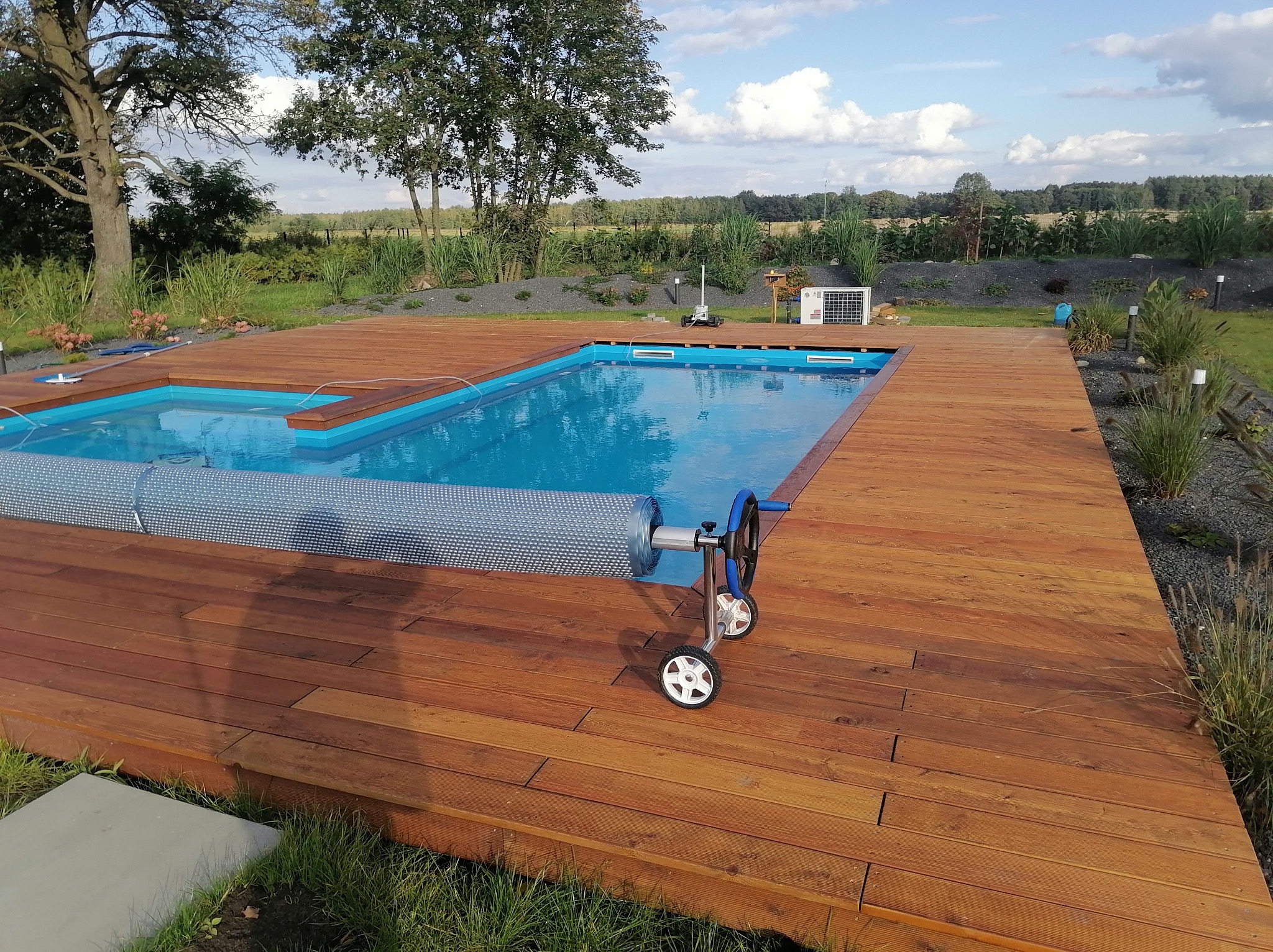 Drewniany taras okalający basen z błękitną wodą, częściowo przykryty roletą. W tle trawa, drzewa i krzewy, co tworzy naturalne otoczenie.