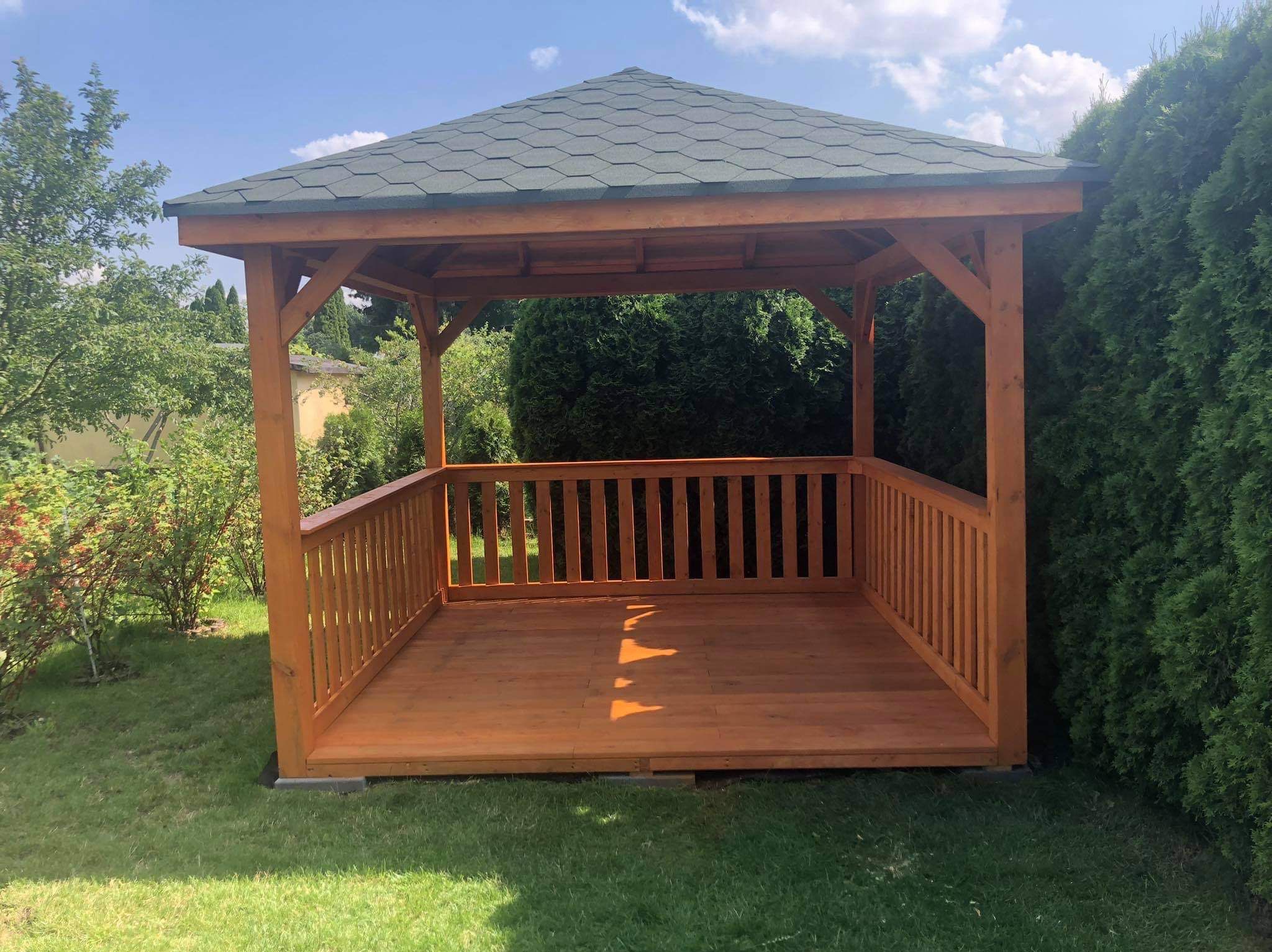 Drewniana altana z zielonym dachem w ogrodzie, otoczona zielenią. Widoczne drewniane balustrady i podłoga. Słoneczny dzień, bujna roślinność w tle.