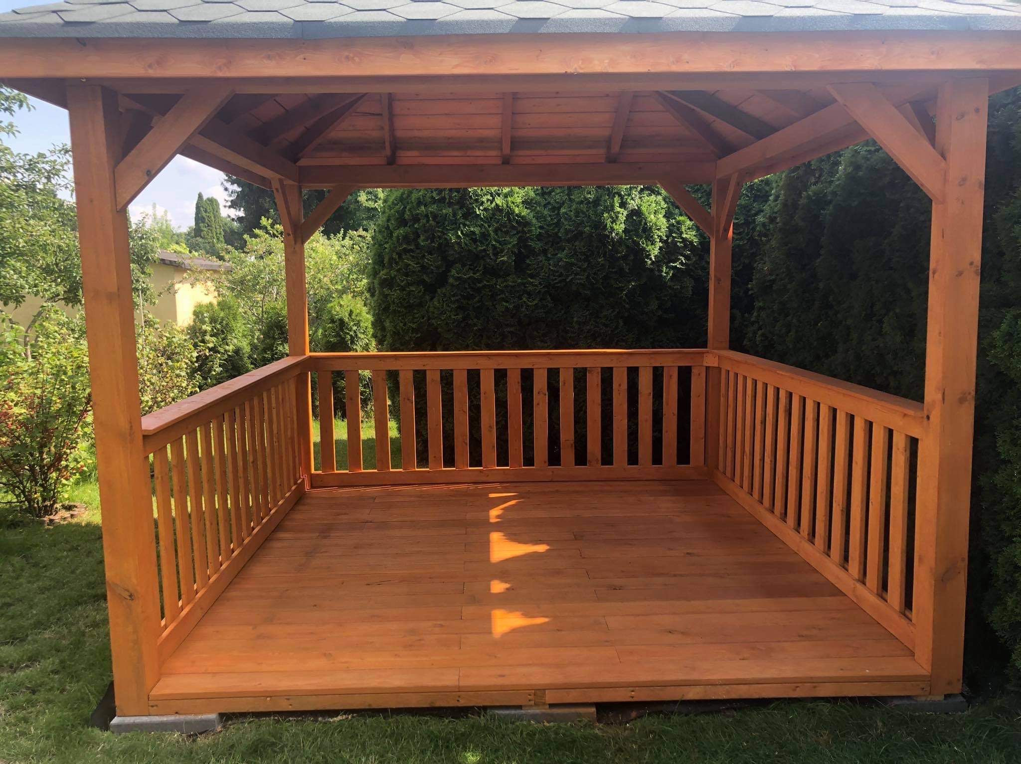 Drewniana altana z balustradą w Wygodzie, zadaszona gontem bitumicznym, otoczona zielenią. Widok z wnętrza, słoneczny dzień.