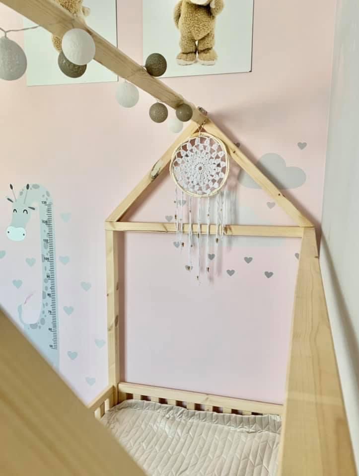 Drewniana konstrukcja łóżka w kształcie domku w pokoju dziecka, ozdobiona łapaczem snów i girlandą świetlną, z grafiką żyrafy i pluszowego misia na ścianie.