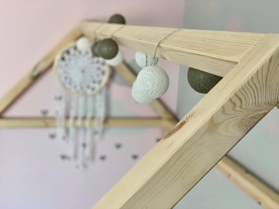 Drewniana konstrukcja w kształcie domku dziecięcego, z zawieszkami w postaci kul i łapacza snów. Jasne drewno, delikatne oświetlenie, pastelowe tło.