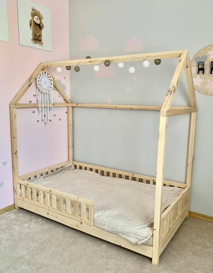 Drewniane łóżko domek dla dziecka z barierkami, ozdobione girlandą świetlną i łapaczem snów, ustawione w pokoju z pastelowymi ścianami i dekoracjami.