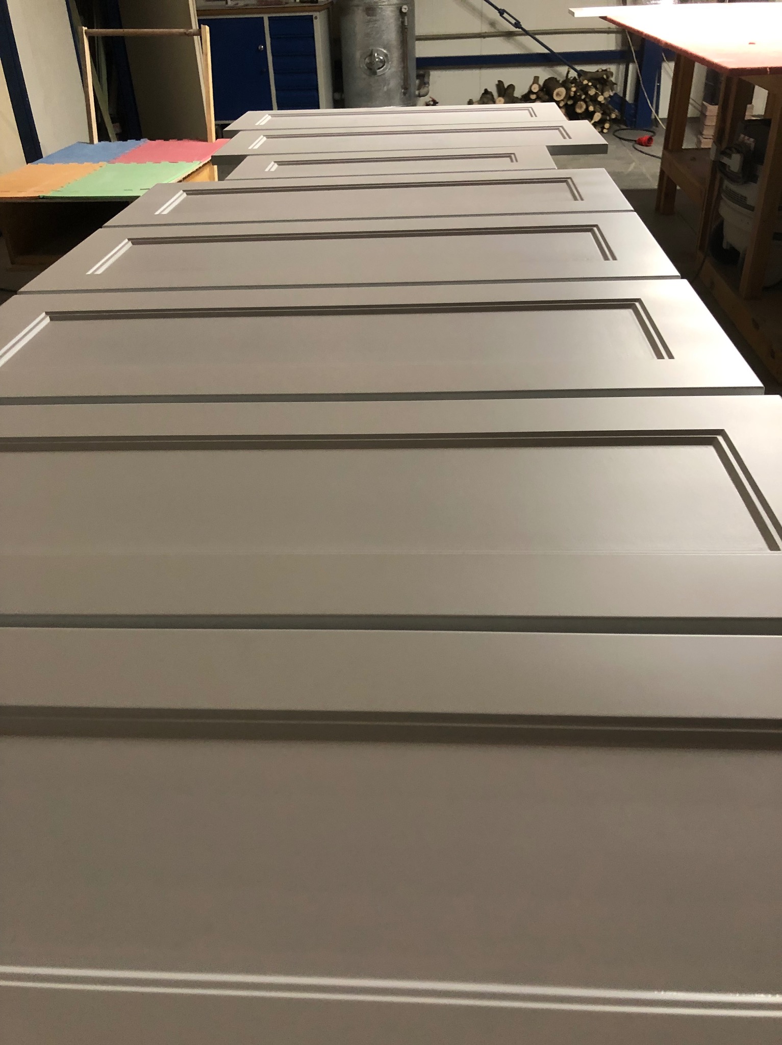 Szare, gotowe fronty meblowe z frezowanymi ramkami leżące obok siebie w warsztacie stolarskim, przygotowane do montażu. Widoczne tło warsztatowe.