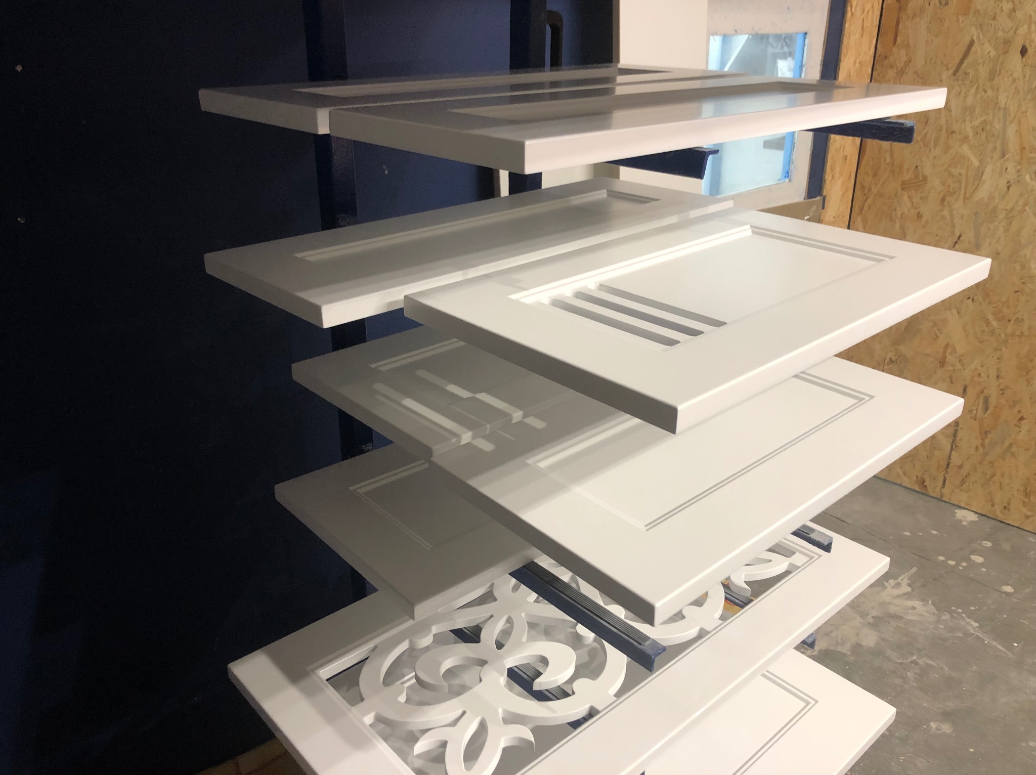 Białe, lakierowane fronty meblowe z różnymi wzorami, ułożone warstwowo na metalowym stojaku w lakierni. Widoczne detale wykończenia i struktura drewna.