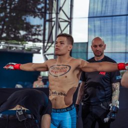 Grzegorz Kowalski - Zawodnik MMA z rozłożonymi ramionami przed walką, widoczny tatuaż z logotypem sponsora na torsie, w tle ring, oświetlenie sceniczne i osoby z obsługi.
