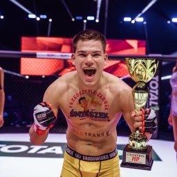 Grzegorz Kowalski - Zwycięski zawodnik sportów walki z pucharem, w ringu, otoczony przez ring girls. Na ciele widoczne reklamy sponsorów. Emocje, triumf, walka.