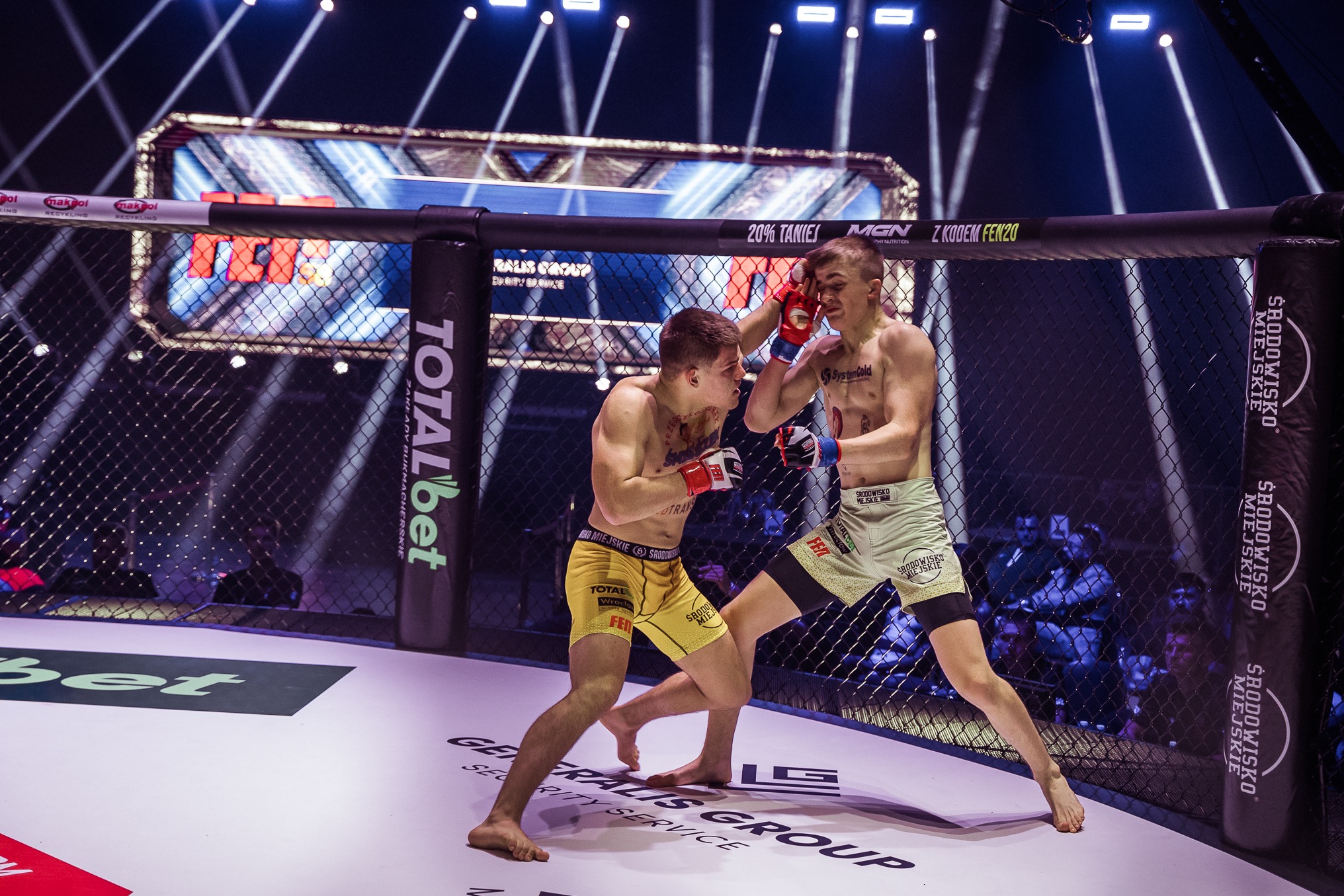 Dwóch zawodników MMA w ringu podczas walki, w tle widownia i oświetlenie. Jeden zawodnik w żółtych spodenkach, drugi w jasnych. Dynamiczne ujęcie.