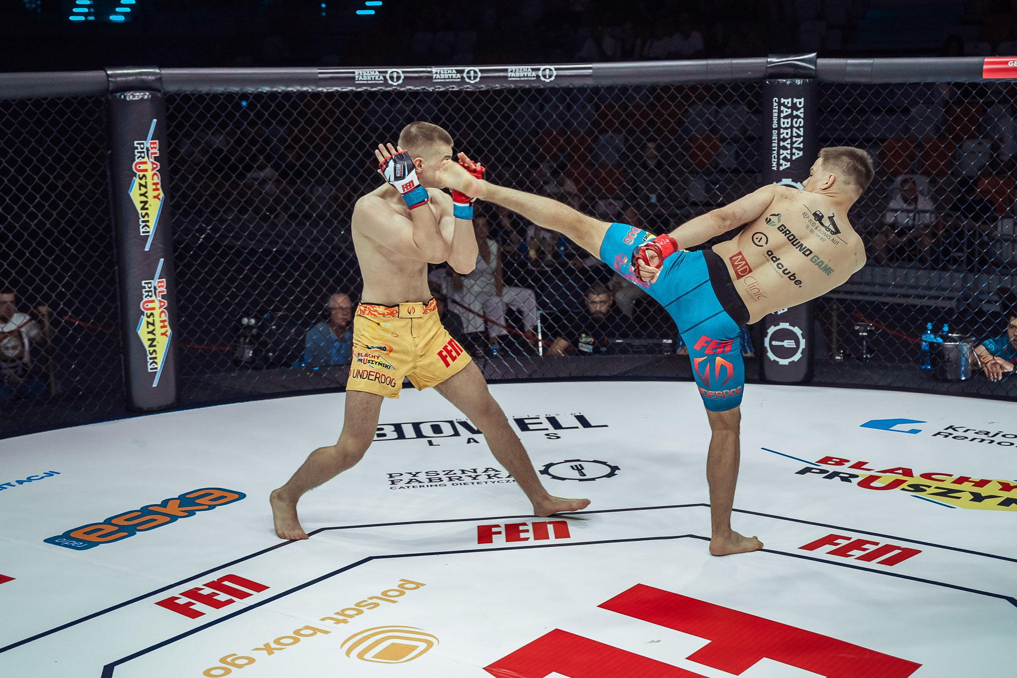 Dynamiczne ujęcie z walki MMA: zawodnik w niebieskich spodenkach wykonuje kopnięcie w kierunku przeciwnika w żółtych spodenkach na ringu z logotypami sponsorów.