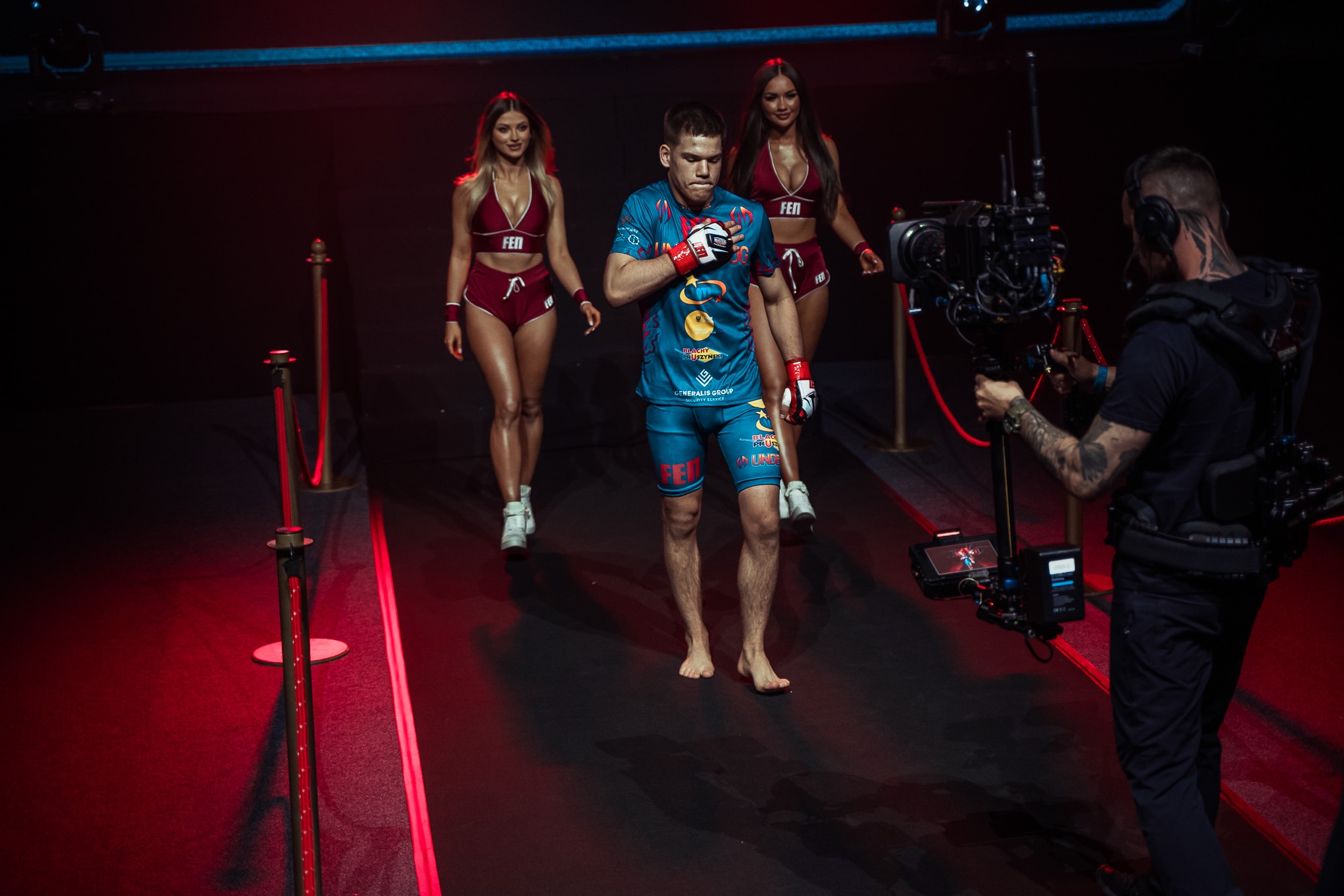 Zawodnik MMA w ringu, otoczony przez ring girls, filmowany przez operatora z kamerą na stabilizatorze. Intensywne oświetlenie sceniczne.