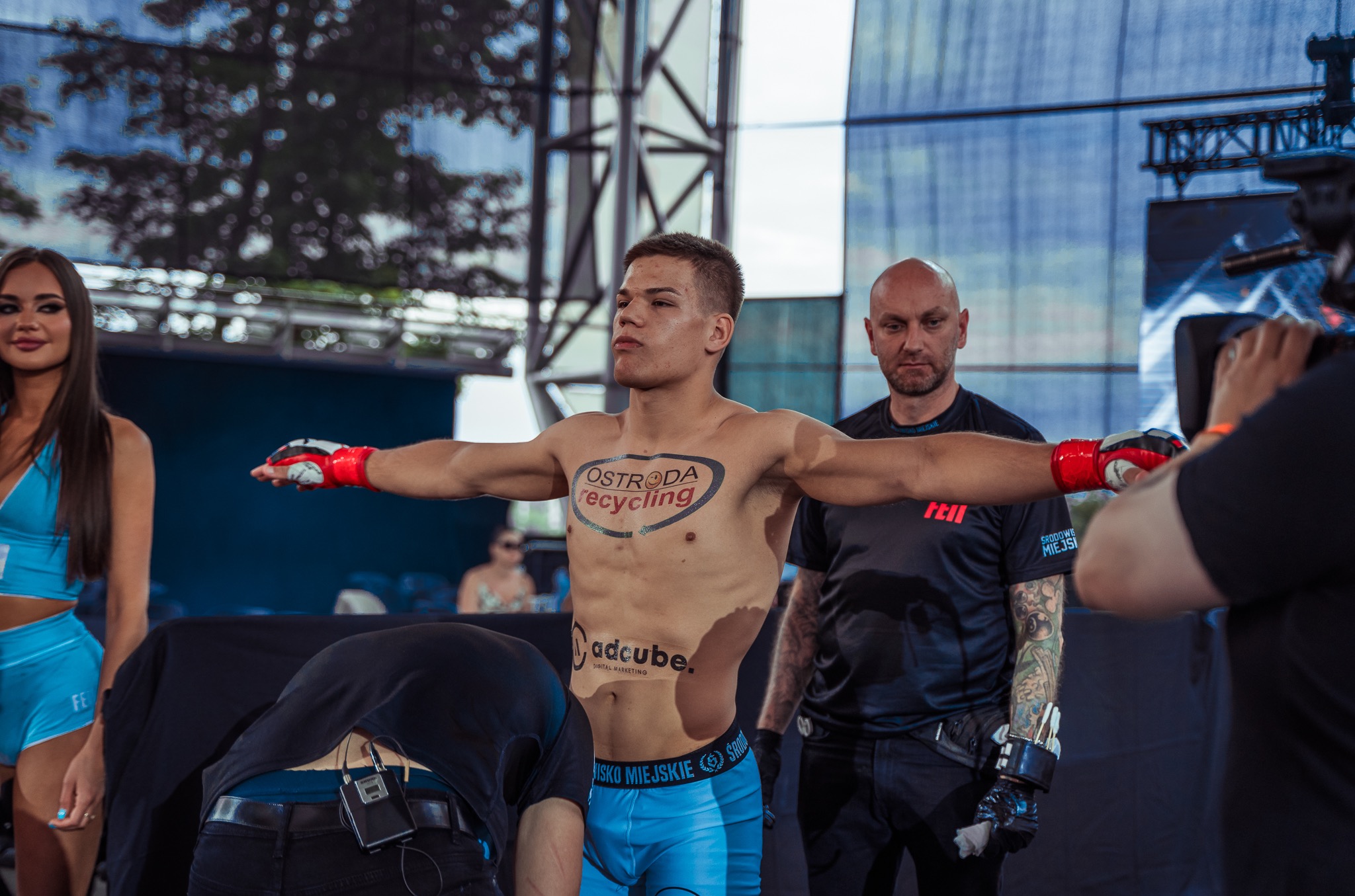 Zawodnik MMA z rozłożonymi ramionami przed walką, widoczny tatuaż z logotypem sponsora na torsie, w tle ring, oświetlenie sceniczne i osoby z obsługi.