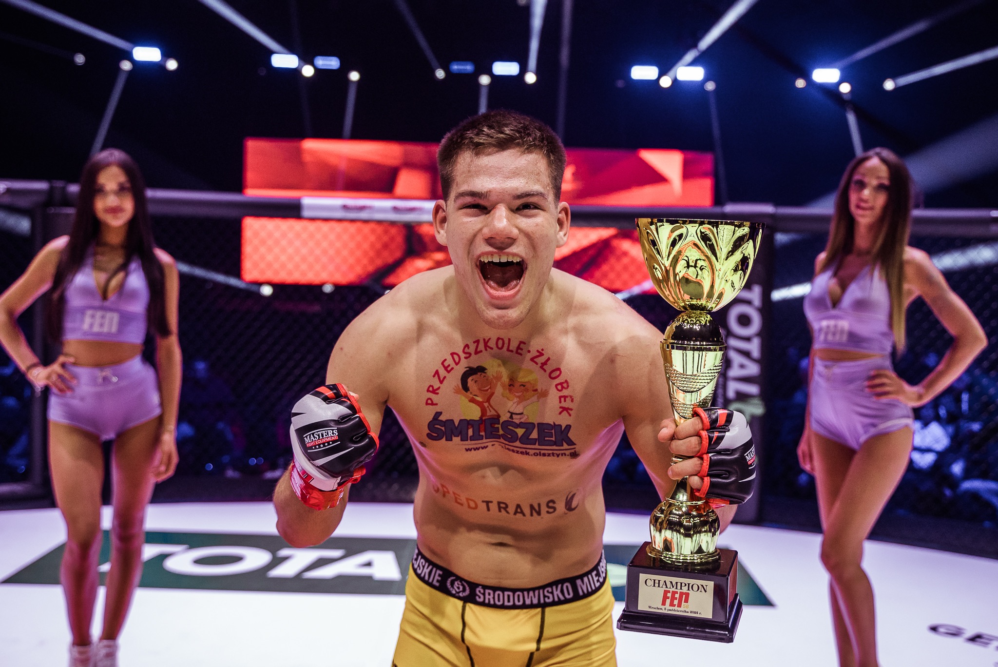 Zwycięski zawodnik sportów walki z pucharem, w ringu, otoczony przez ring girls. Na ciele widoczne reklamy sponsorów. Emocje, triumf, walka.
