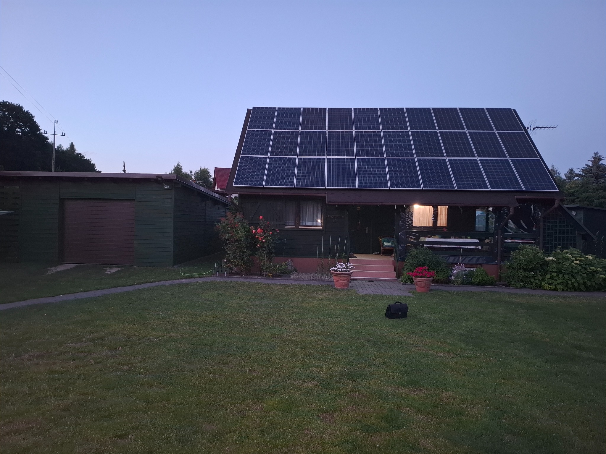 Dom jednorodzinny z panelami fotowoltaicznymi na dachu, widok z ogrodu, wieczorne oświetlenie. Dodatkowo widoczny garaż i zadbany trawnik.