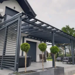 Nowoczesna pergola metalowa w kolorze grafitowym przylegająca do domu z jacuzzi i meblami ogrodowymi. Uwagę zwracają trzy drzewka w donicach.