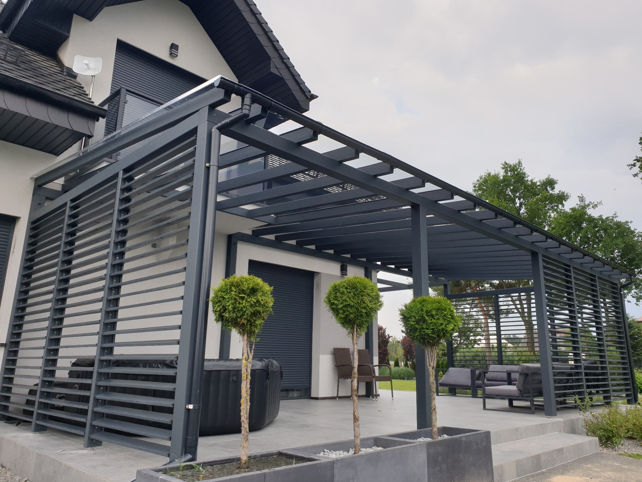 Nowoczesna pergola metalowa w kolorze grafitowym przylegająca do domu z jacuzzi i meblami ogrodowymi. Uwagę zwracają trzy drzewka w donicach.