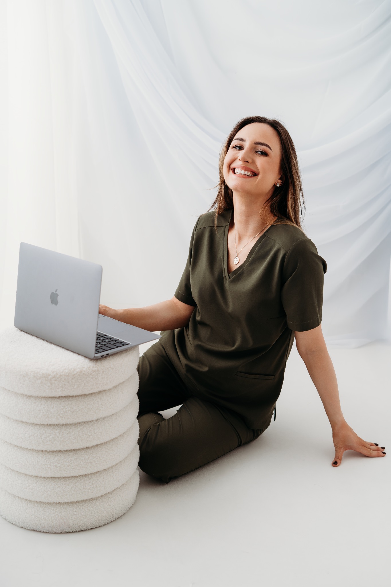 Uśmiechnięta kobieta w zielonym uniformie siedzi na podłodze z laptopem na pufie. Jasne tło, naturalne światło, widoczna biżuteria i ciemny manicure.