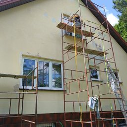 BUD-MAR - Pracownik na rusztowaniu szpachluje elewację domu z widocznymi ubytkami tynku, przygotowując ją do malowania. Widoczne okna, dach i fragment zieleni.