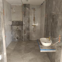 BUD-MAR - Łazienka w trakcie wykańczania: szare płytki imitujące marmur, złota armatura, podwieszana umywalka. Widoczne kable i taśma malarska.