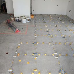 BUD-MAR - Szare płytki podłogowe, częściowo ułożone, z systemem poziomowania. W tle wiaderka z zaprawą i narzędzia glazurnicze. Widoczne kable wystające z otworu w podłodze.