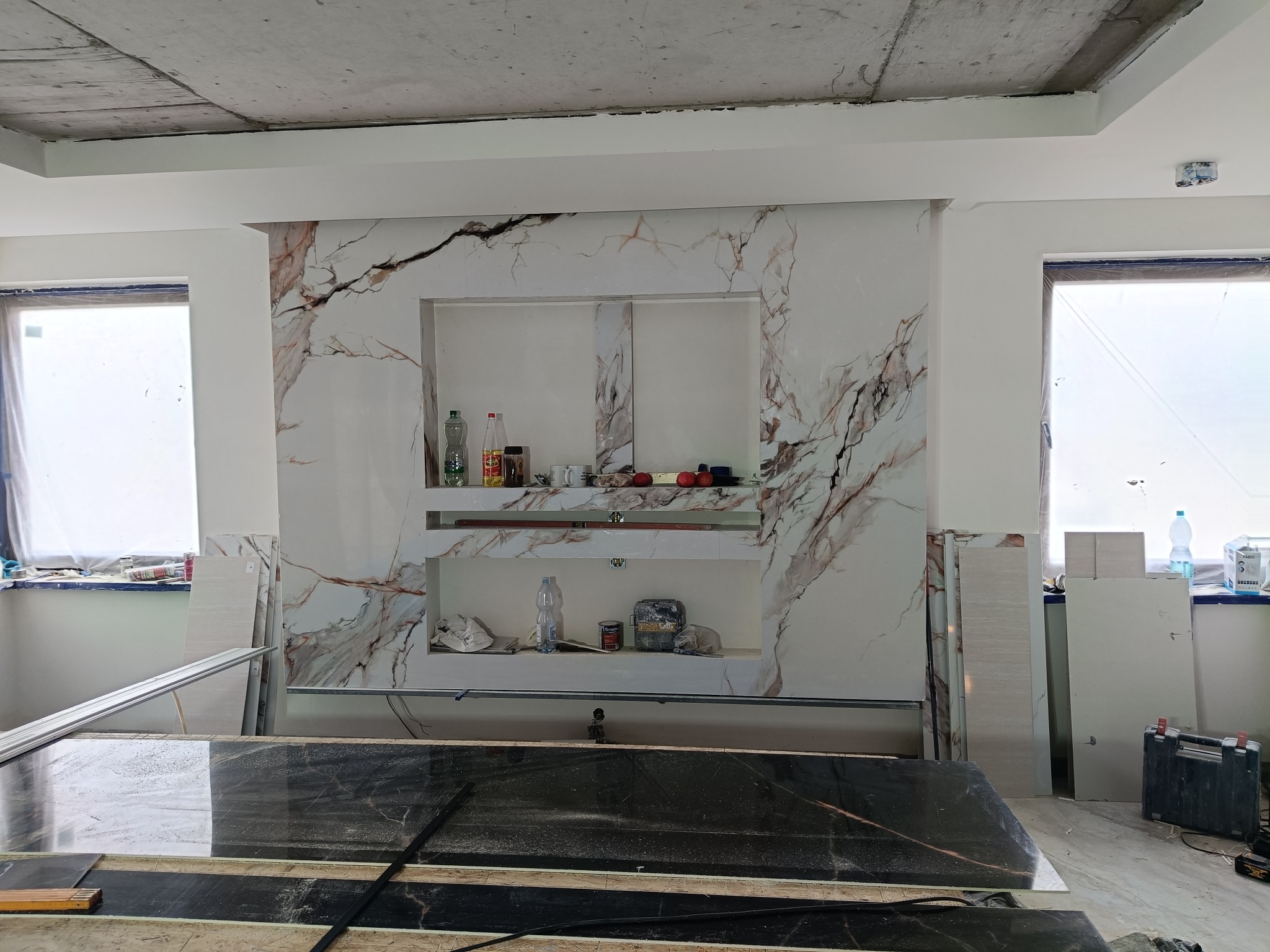 Ściana z płyt imitujących marmur, z wnękami na półki, w trakcie montażu. Widoczne narzędzia i materiały budowlane. Surowy betonowy sufit, okna.