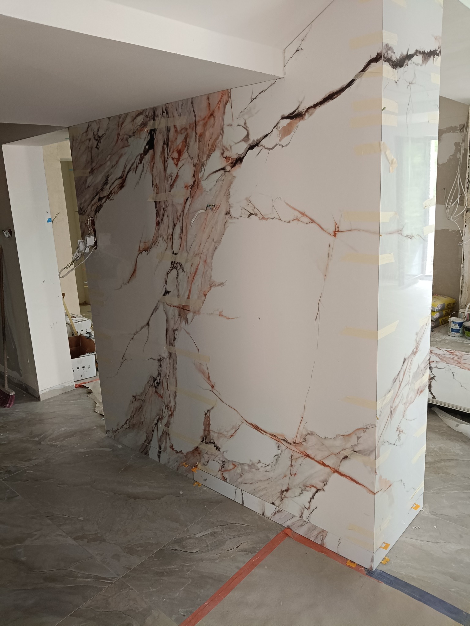 Ściana wykończona płytami imitującymi marmur z widocznymi taśmami mocującymi, w trakcie prac wykończeniowych wnętrza, na szarej podłodze z płytek.