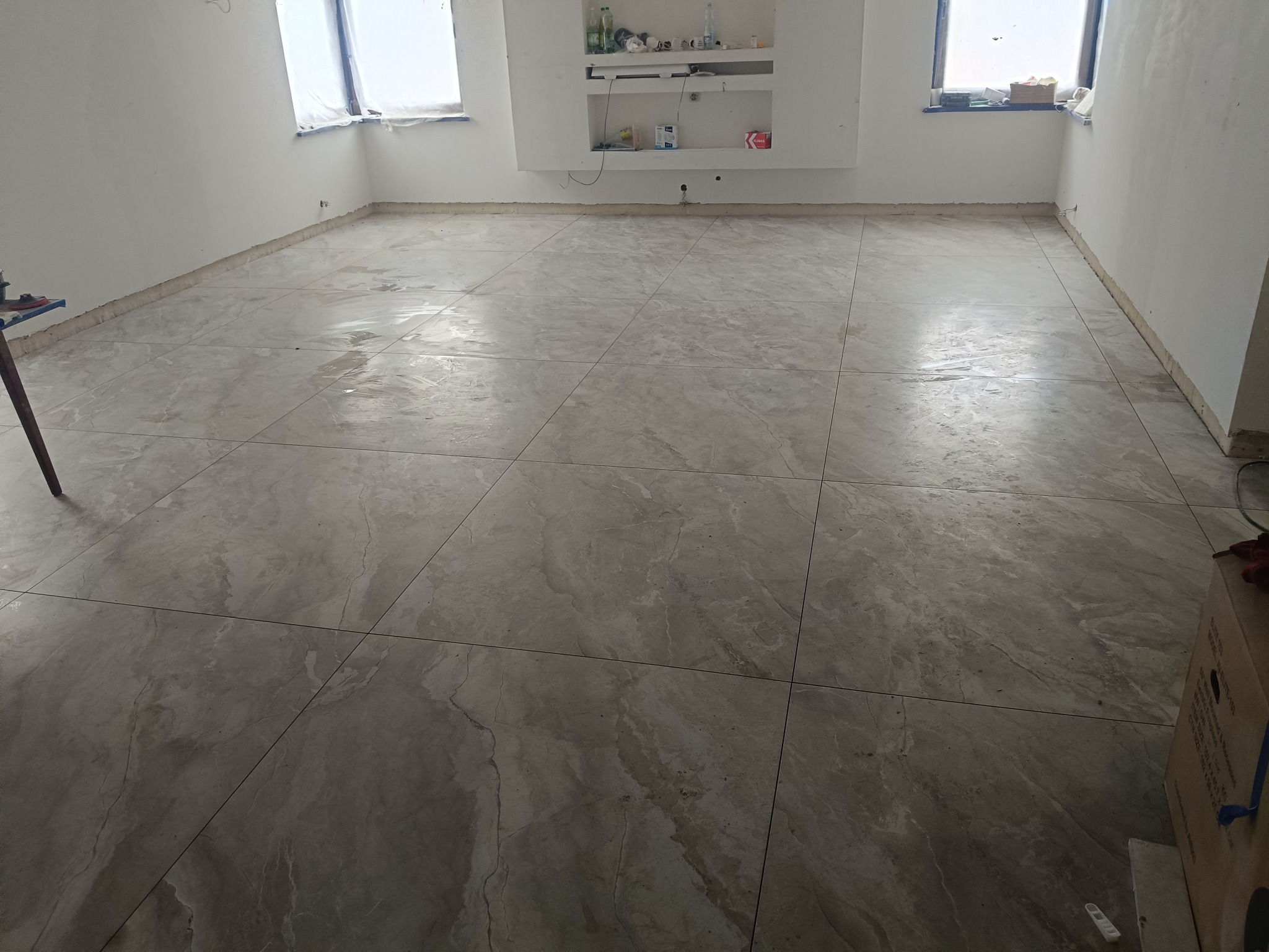 Wykończona podłoga z dużych, szarych płytek w pustym pokoju z oknami. Widoczne fugi i fragmenty narzędzi glazurniczych. Świeżo ułożona podłoga, gotowa do użytku.