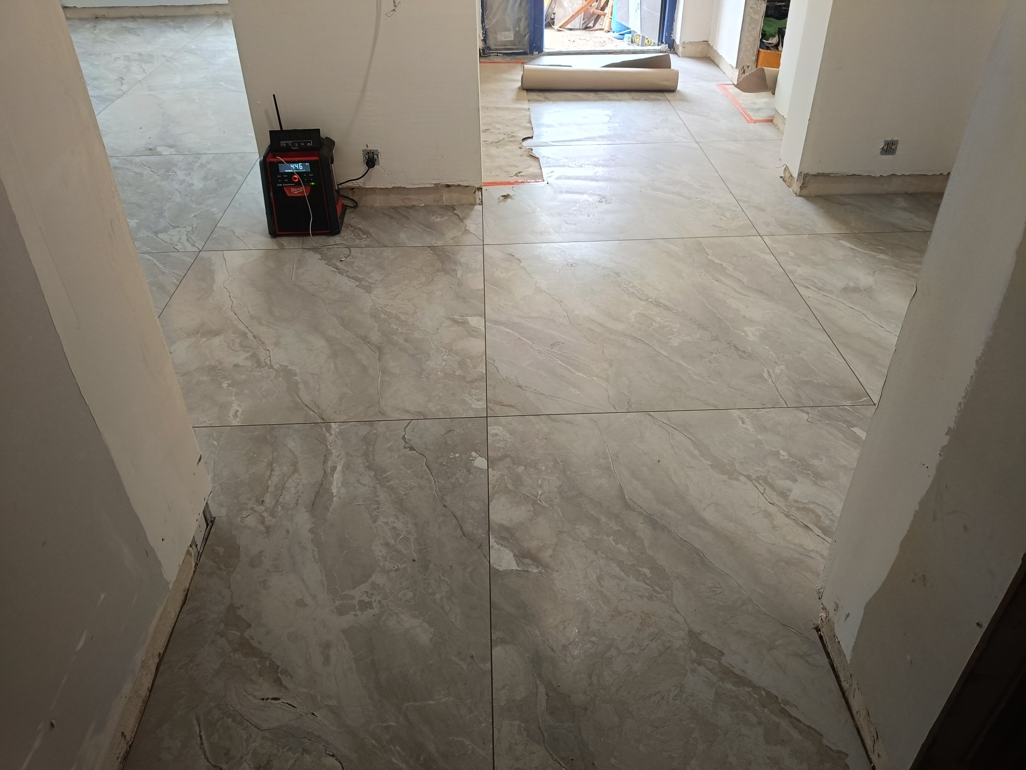 Beżowe płytki podłogowe z wzorem marmuru, ułożone w trakcie remontu. Widoczny sprzęt budowlany przy ścianie i częściowo zabezpieczona podłoga w przejściu.