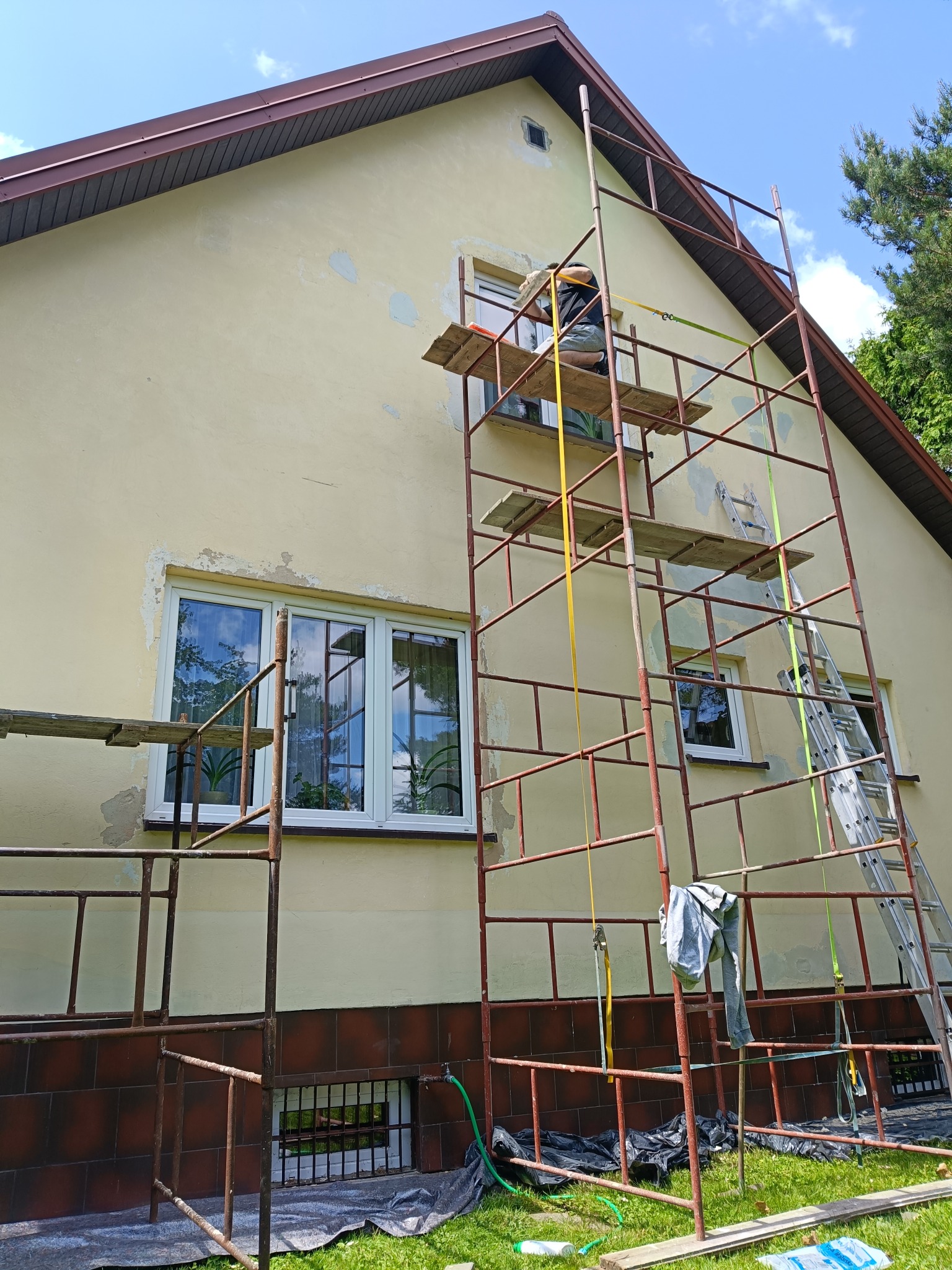 Pracownik na rusztowaniu szpachluje elewację domu z widocznymi ubytkami tynku, przygotowując ją do malowania. Widoczne okna, dach i fragment zieleni.