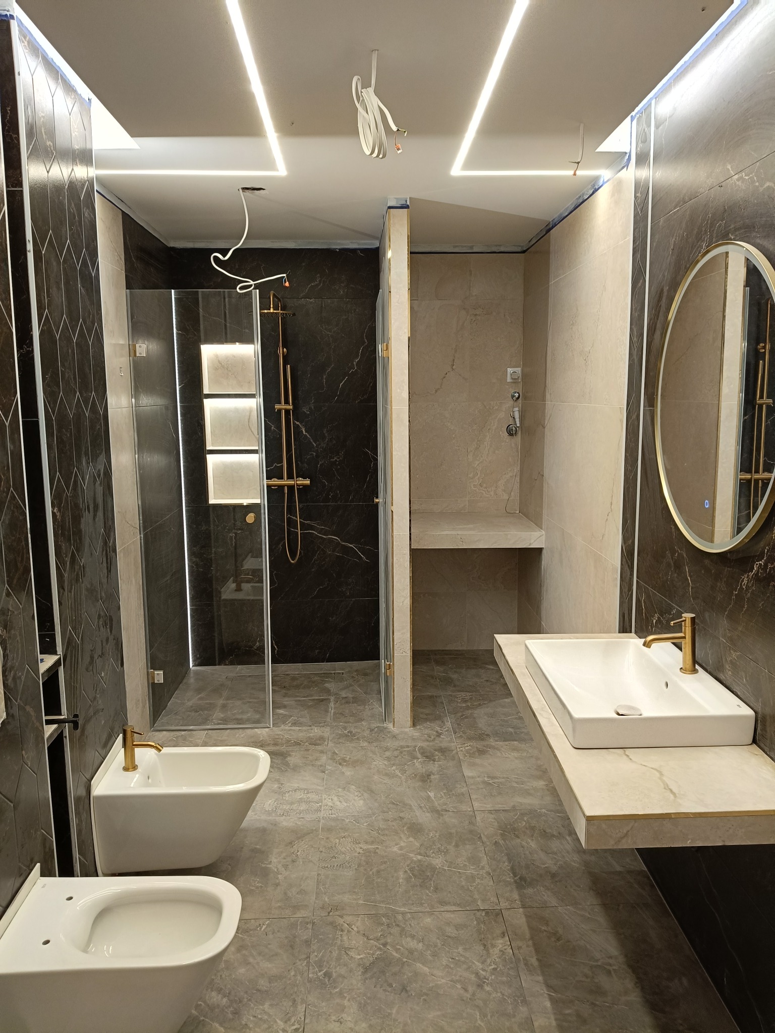 Nowoczesna łazienka z kabiną prysznicową typu walk-in, umywalką na blacie i podświetleniem LED. Ciemne płytki imitujące marmur. Wnętrze w trakcie wykańczania.