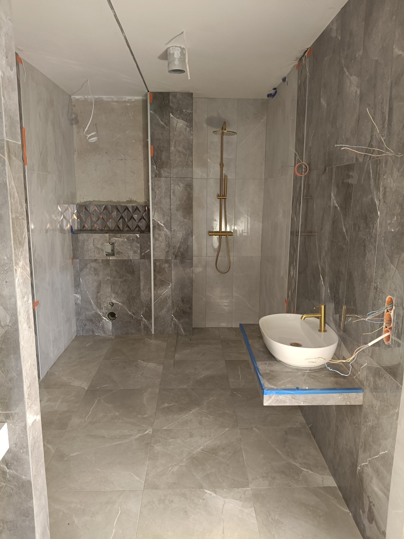 Łazienka w trakcie wykańczania: szare płytki imitujące marmur, złota armatura, podwieszana umywalka. Widoczne kable i taśma malarska.