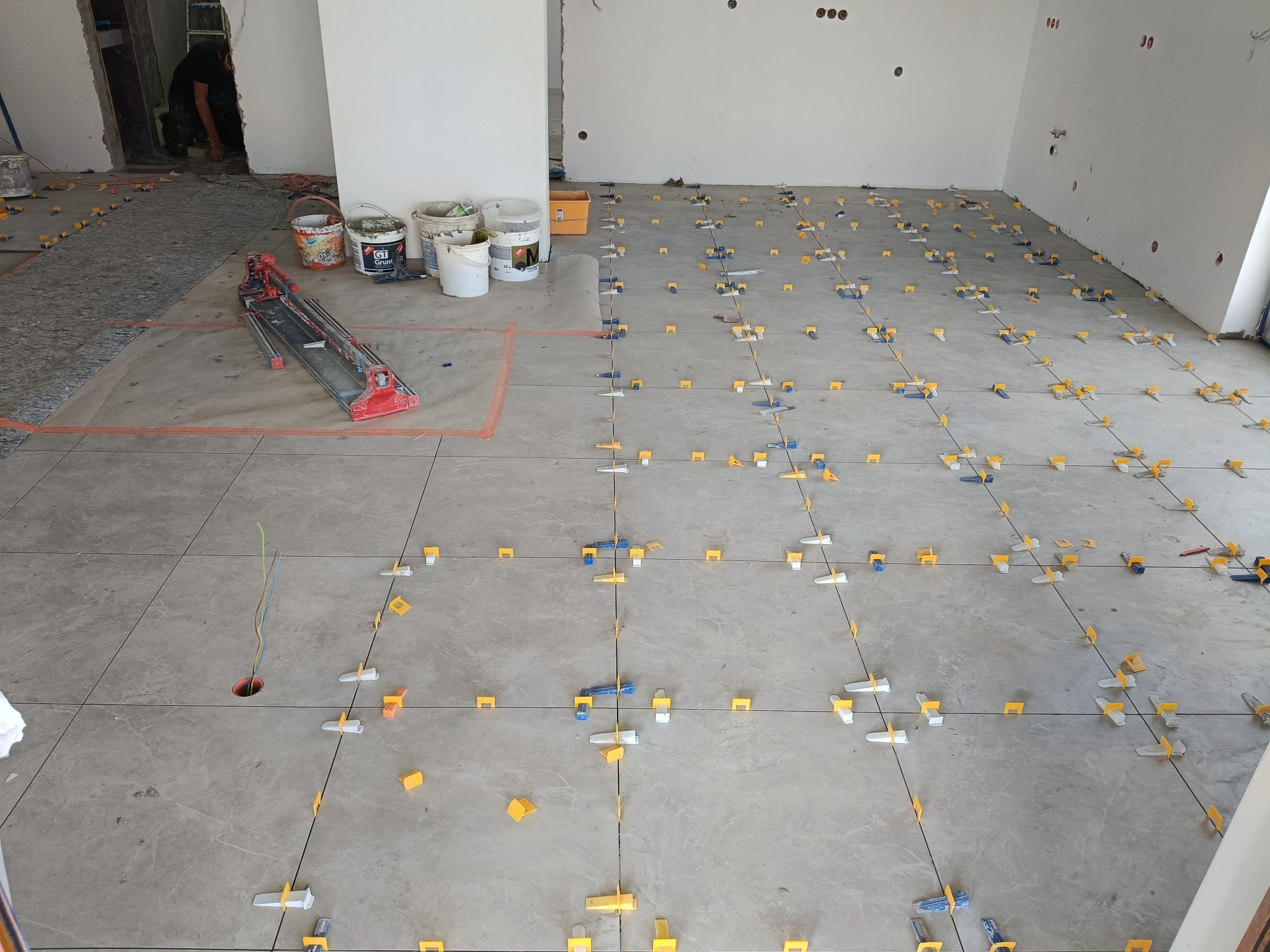 Szare płytki podłogowe, częściowo ułożone, z systemem poziomowania. W tle wiaderka z zaprawą i narzędzia glazurnicze. Widoczne kable wystające z otworu w podłodze.