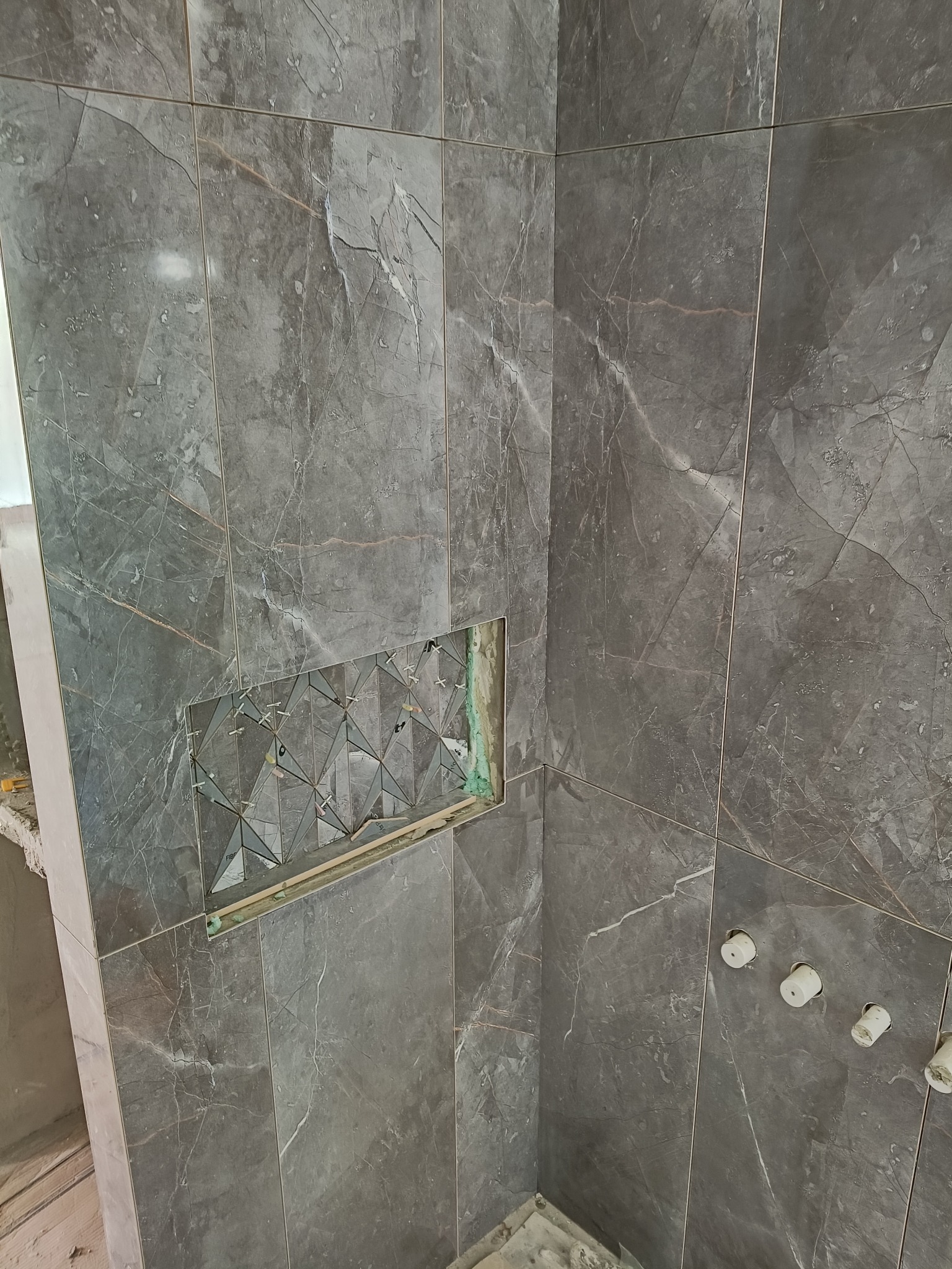 Ściana w łazience z szarymi płytkami imitującymi marmur, z wnęką wyłożoną mozaiką. Widoczne krzyżyki dystansowe i fragment instalacji wodnej.