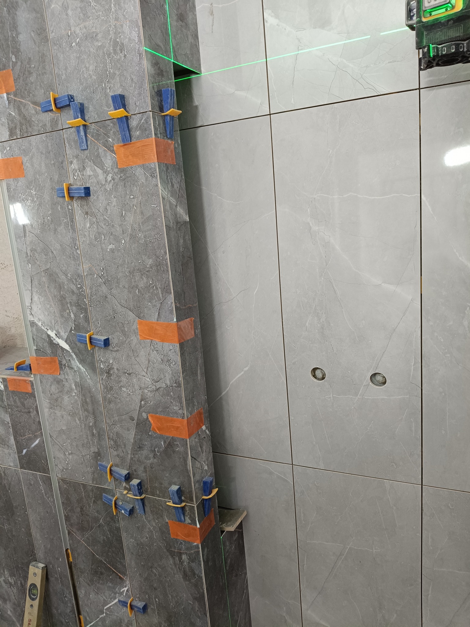 Montaż płytek imitujących marmur na ścianie z użyciem poziomicy laserowej i klipsów poziomujących. Widoczne otwory na gniazdka.