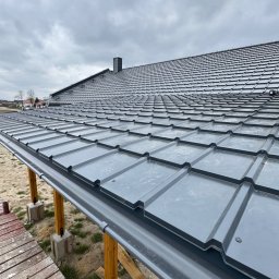 Dekarstwo-Ciesielstwo Patryk Chmielinski - Szare dachówki na nowym dachu domu z zamontowanymi rynnami, widziane z perspektywy rusztowania. Pochmurne niebo w tle.