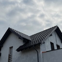 Dekarstwo-Ciesielstwo Patryk Chmielinski - Dom jednorodzinny z nowym, ciemnym dachem z dachówki ceramicznej i rynnami. Widoczne zabezpieczone okna i elewacja w trakcie prac wykończeniowych.